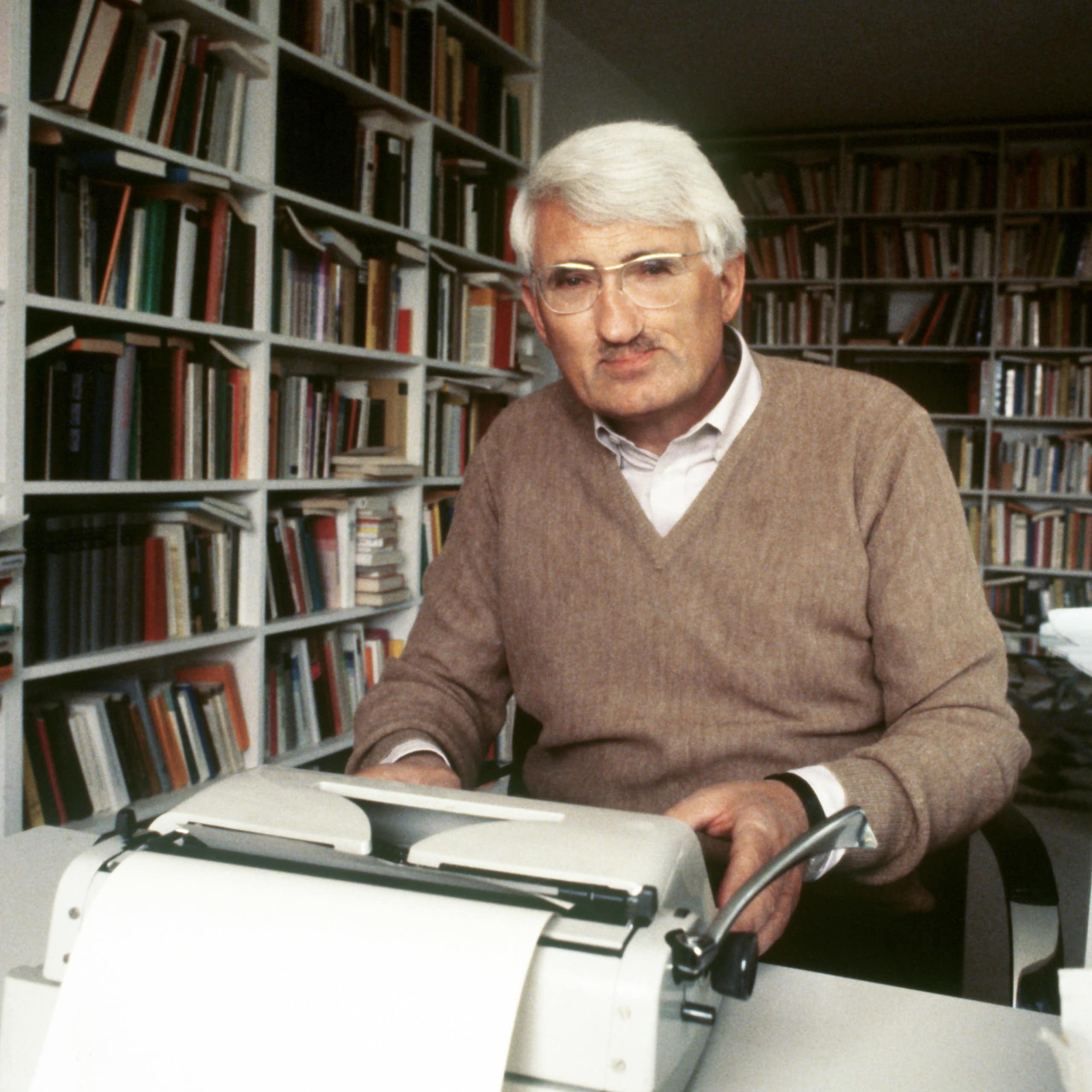 Man at his typewriter in a book-lined room.