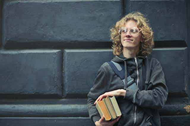 A young man stands against a concrete wall, holding books. 
