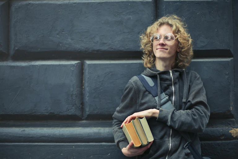 A young man stands against a concrete wall, holding books.