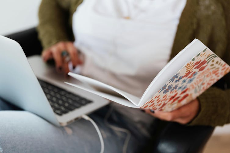 A young woman looks at a notebook with a floral cover and types on a laptop.