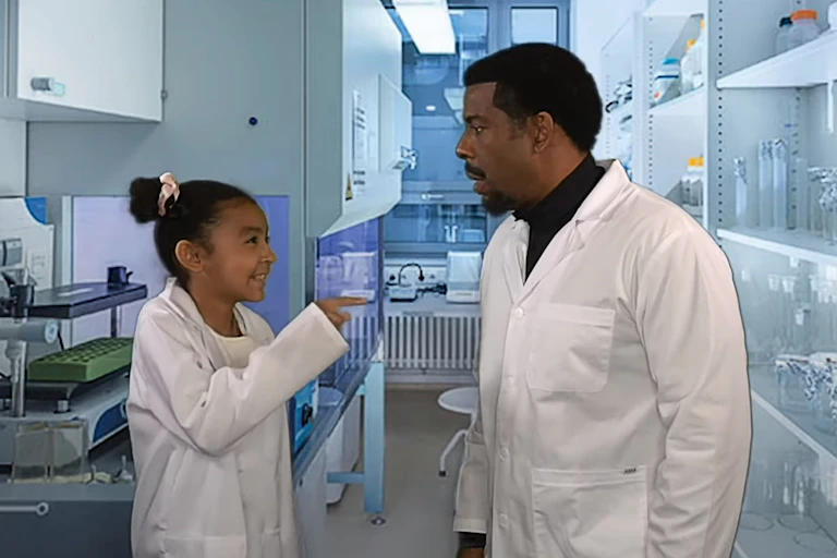 A young girl grins as she points her finger at her dad. Both are wearing scientists' lab coats in a lab setting.