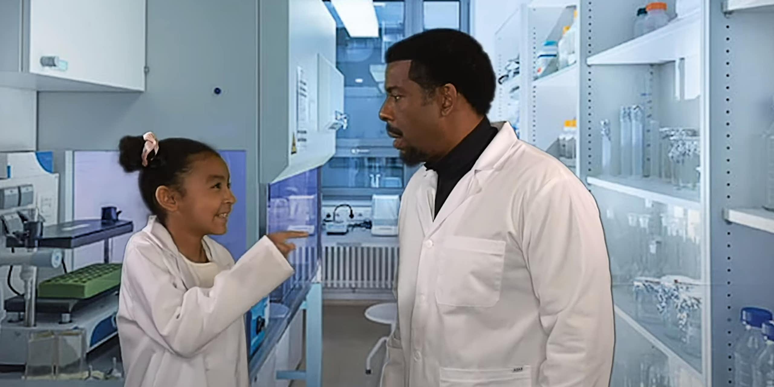 A young girl grins as she points her finger at her dad. Both are wearing scientists' lab coats in a lab setting.