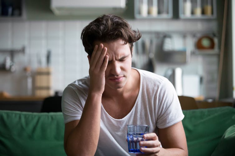 A dehydrated man with a headache holds his head in pain while holding a glass of water.