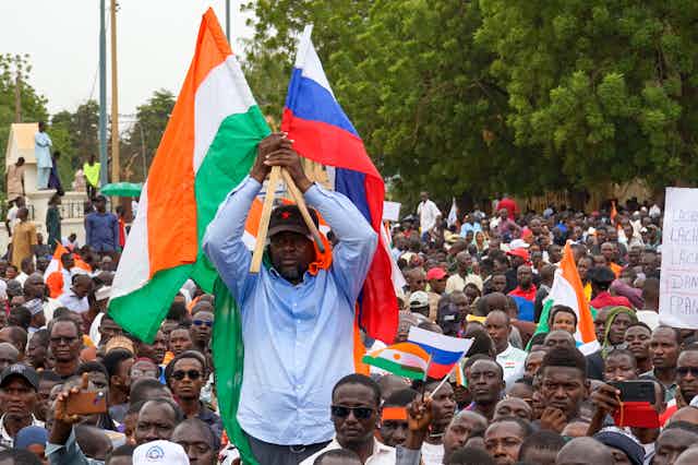 An African man holding the flags of Niger and Russia being held aloft above a large crowd.