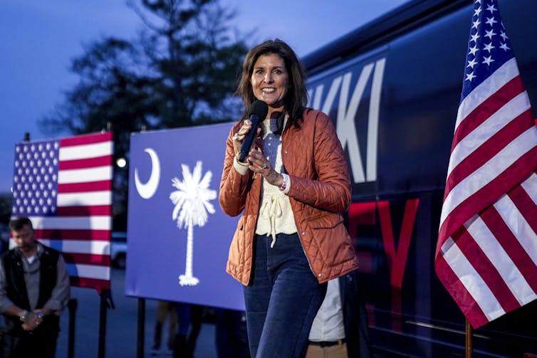 Una donna sta su un palco con in mano un microfono davanti a una bandiera degli Stati Uniti e alla bandiera dello stato della Carolina del Sud.