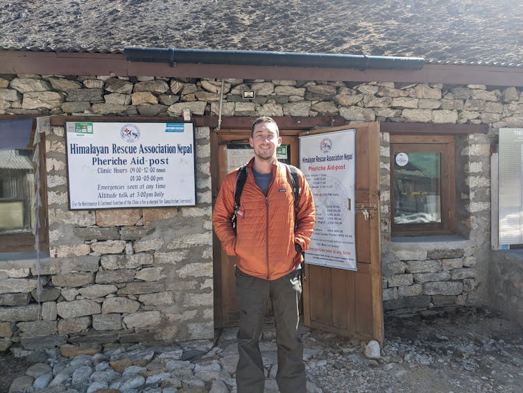 Researcher wearing cold-weather gear stands in front of a health clinic in the Himalayas.
