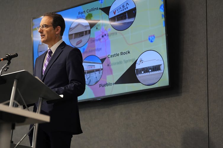 Man in suit and tie stands at a podium in front of a map with photos of supermarkets on it.