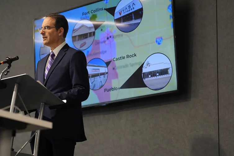 Man in suit and tie stands at a podium in front of a map with photos of supermarkets on it.
