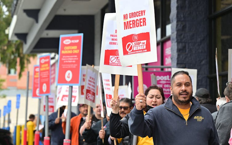 A group of people protest while holding red and white anti-merger pro-labor signs aloft.