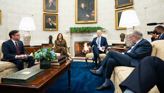 3 men and 1 woman in suits are sitting on couches in an ornate room with presidential portraits on the wall and a fireplace