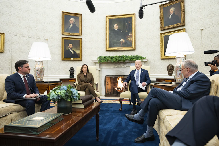 3 men and 1 woman in suits are sitting on couches in an ornate room with presidential portraits on the wall and a fireplace