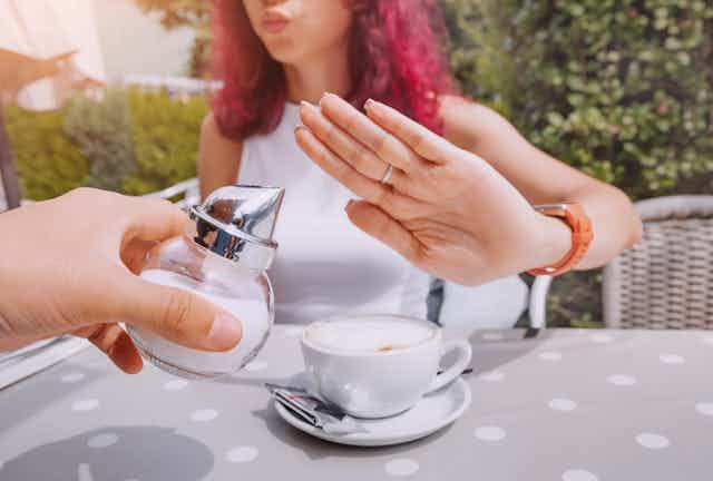Mesa de café com pessoa oferecendo colocar açúcar em bebida e mulher recusando com um gesto