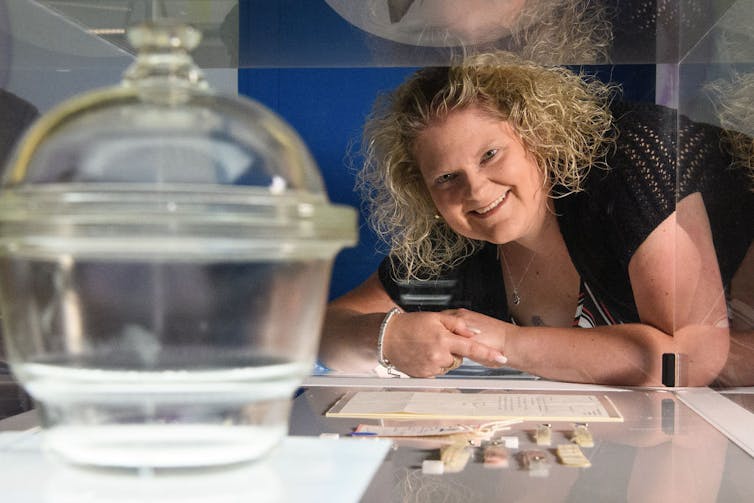 Smiling person with blonde, wavy hair leaning on arms looking at a glass container in an enhibit