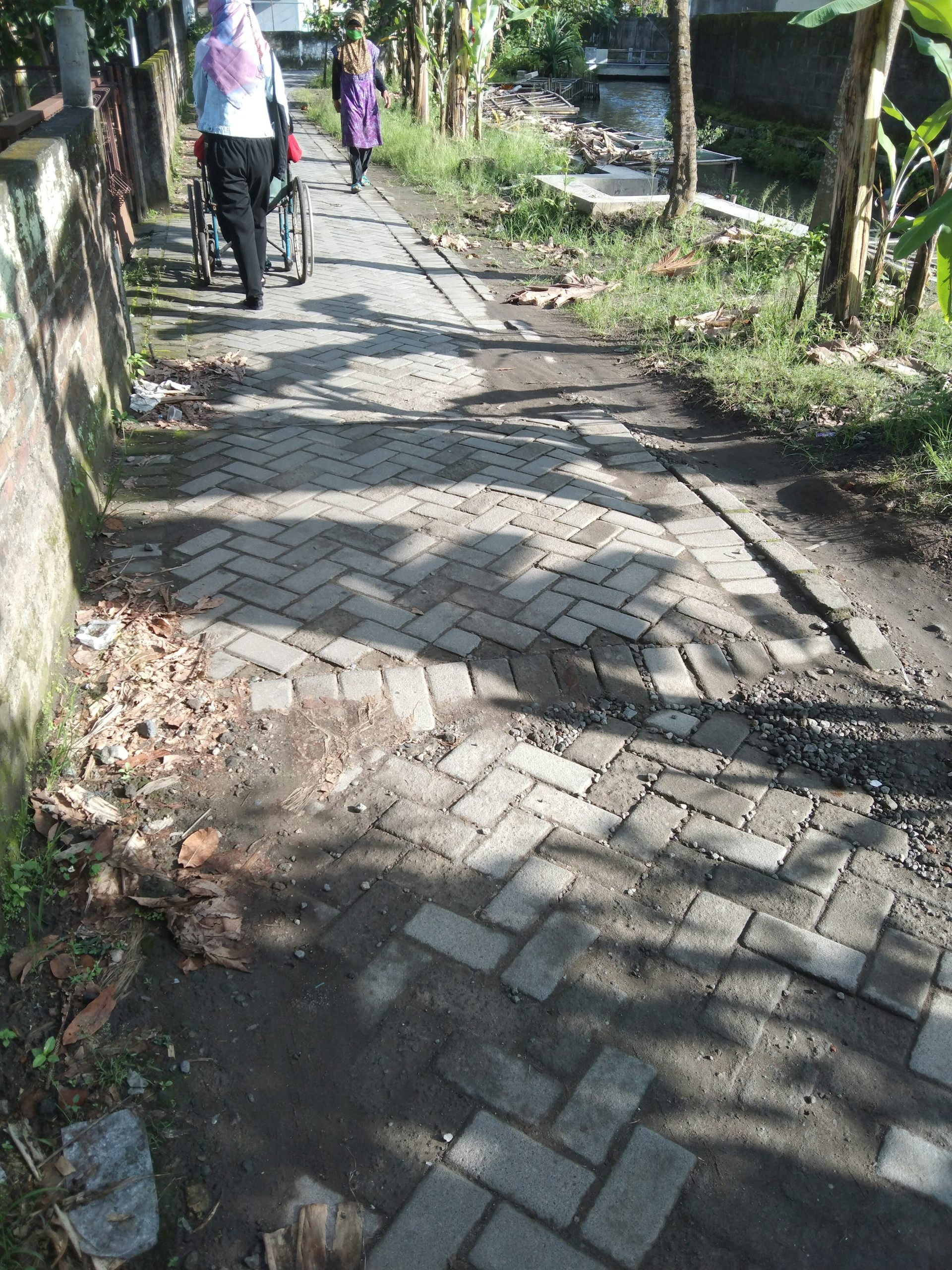 Gang kampung kota di tepi sungai yang permukaannya terbuat dari paving block.