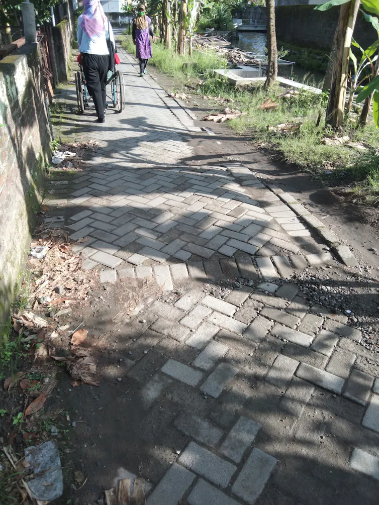 Gang kampung kota di tepi sungai yang permukaannya terbuat dari paving block.