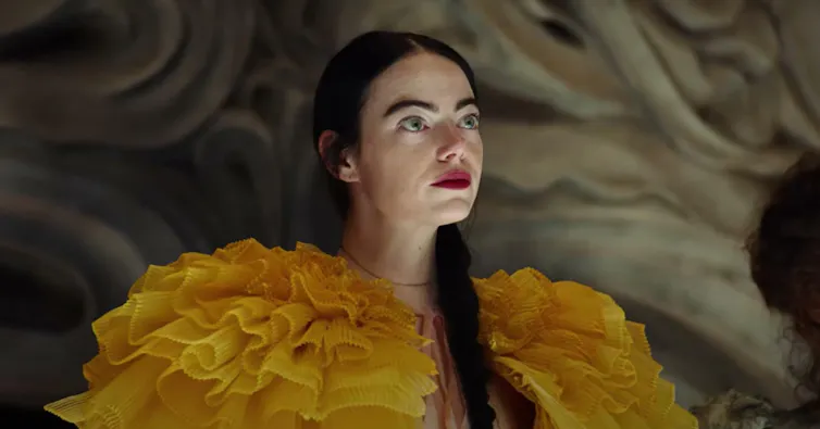 Una mujer vestida con un traje amarillo y con los labios rojos mira al horizonte.