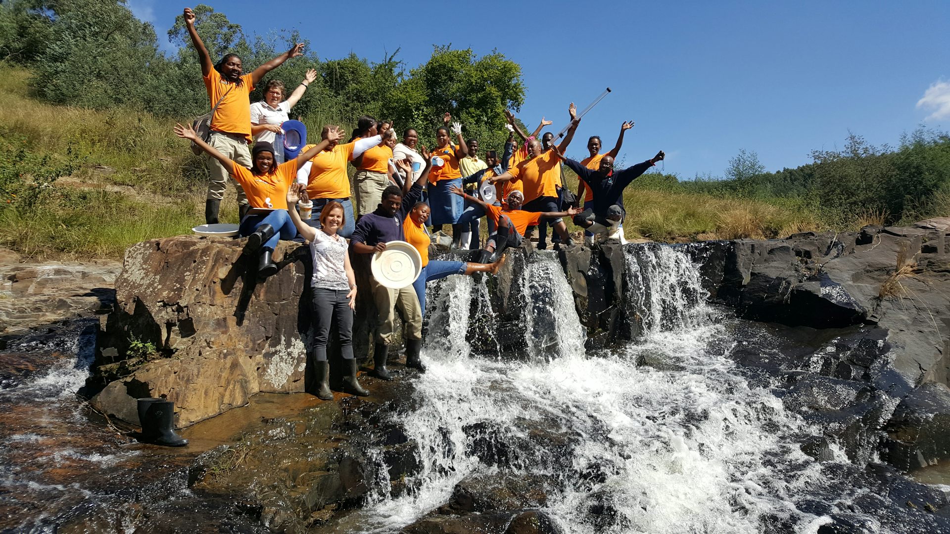 A group of people stand cheering on the bank of a river and looking very happy