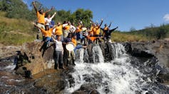 A group of people stand cheering on the bank of a river and looking very happy