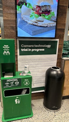 A poster at a supermarket that says camera technology trial in progress, partially obscured by a couple of bins.