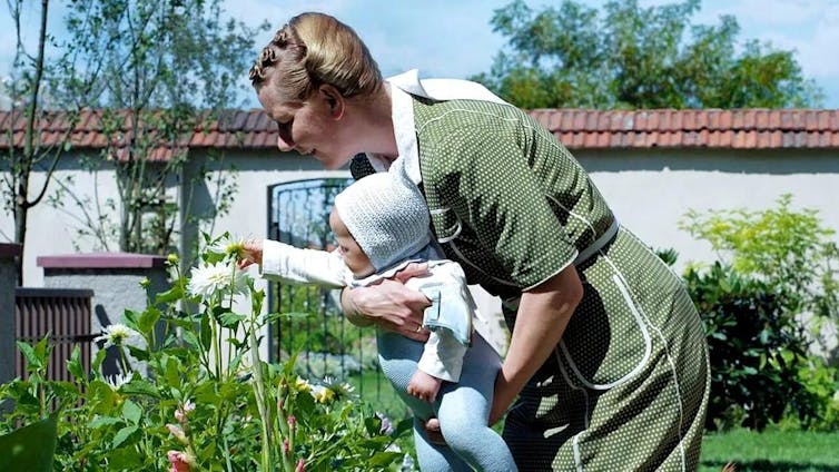 Una mujer observa unas flores mientras se las enseña a un bebé que lleva en brazos.