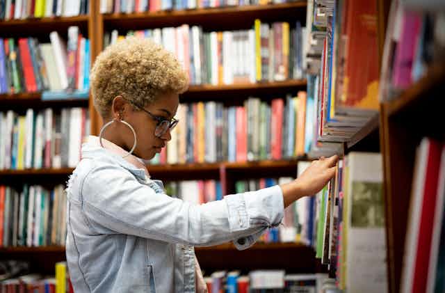 A high school-age girl looks at a book on a rack at the library.