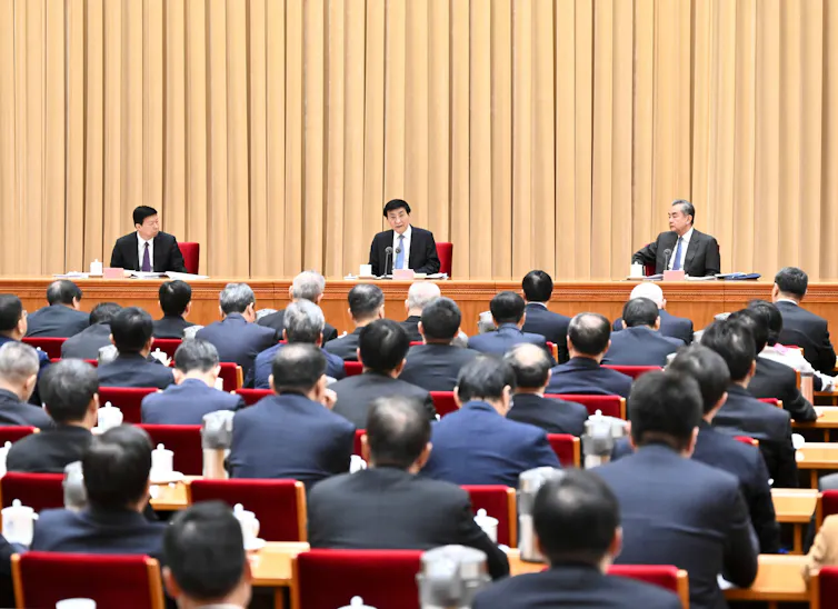 Chinese communist party officials sit on a bench overlooking a hall full of Communist Party delegates.