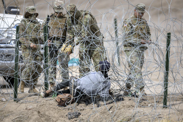 People in camoflauge army outfits stand close to barbed wire in a desert. A person lies on the ground in the middle of the barbed wire.