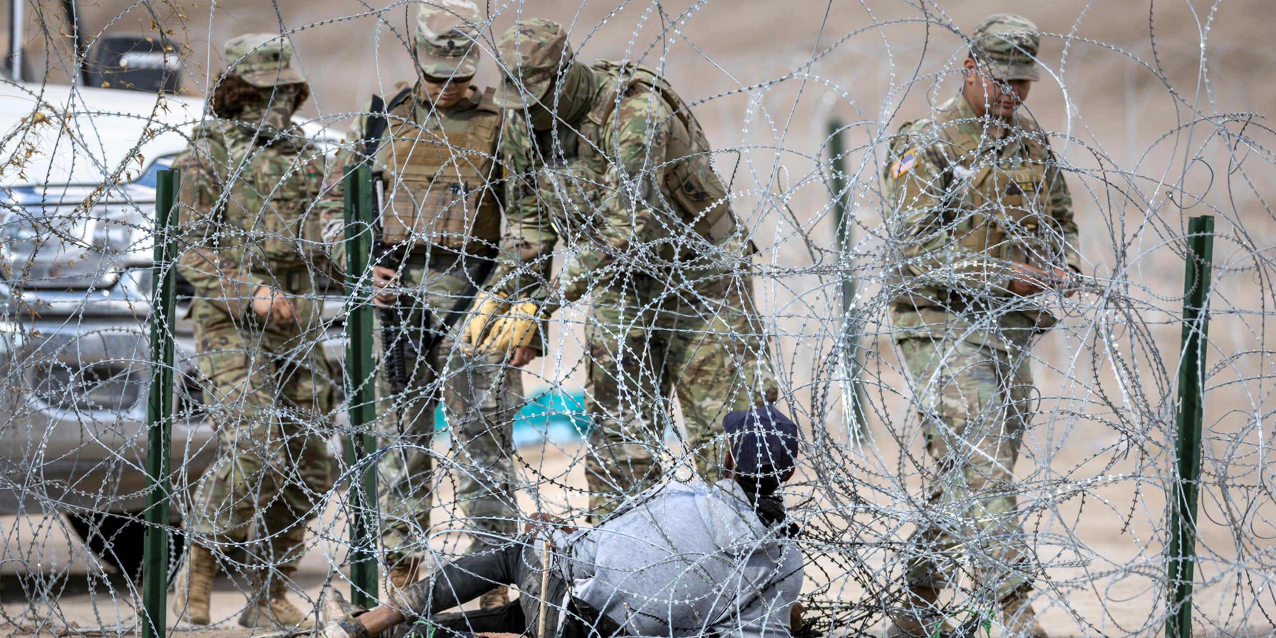 People in camoflauge army outfits stand close to barbed wire in a desert. A person lies on the ground in the middle of the barbed wire. 