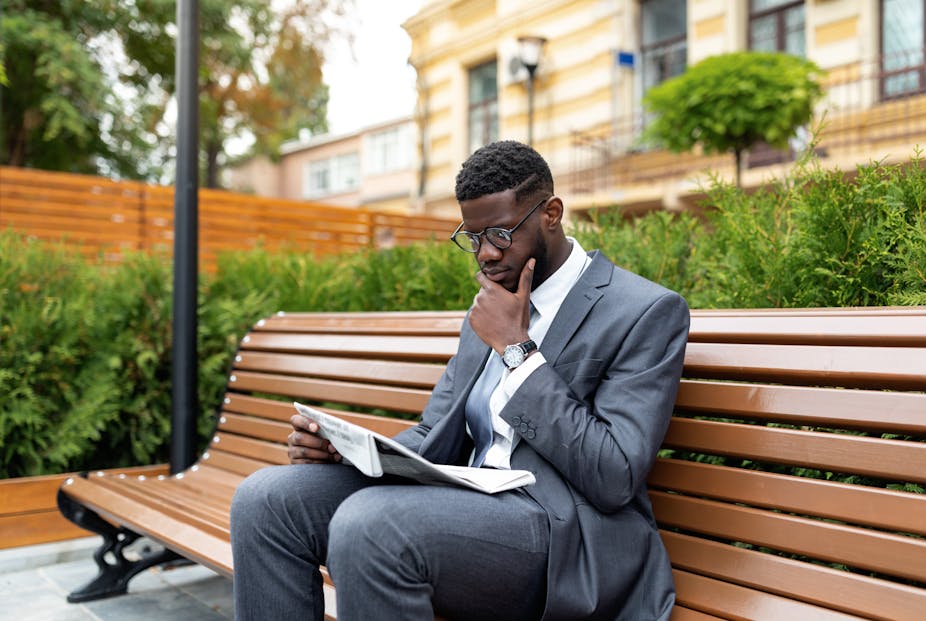 Black businessman in suit reading newspaper while sitting on bench outside