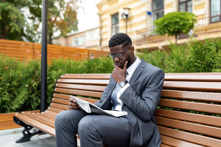 Black businessman in suit reading newspaper while sitting on bench outside