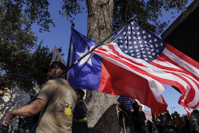 Un hombre es visto de perfil, sosteniendo tanto una bandera estadounidense como una de Texas, que es roja y azul con una estrella blanca.
