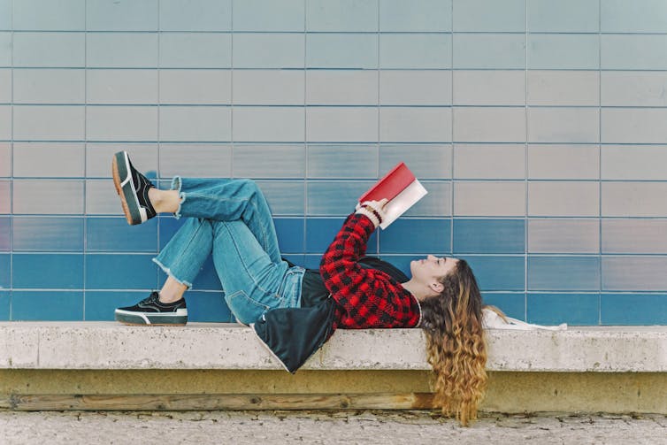 A girl lies on her back on a bench reading a book that she is holding.