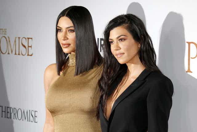 Kim Kardashian wearing a gold, sleeveless dress poses next to Kourtney Kardashian, who wears a black blazer, at an event. 