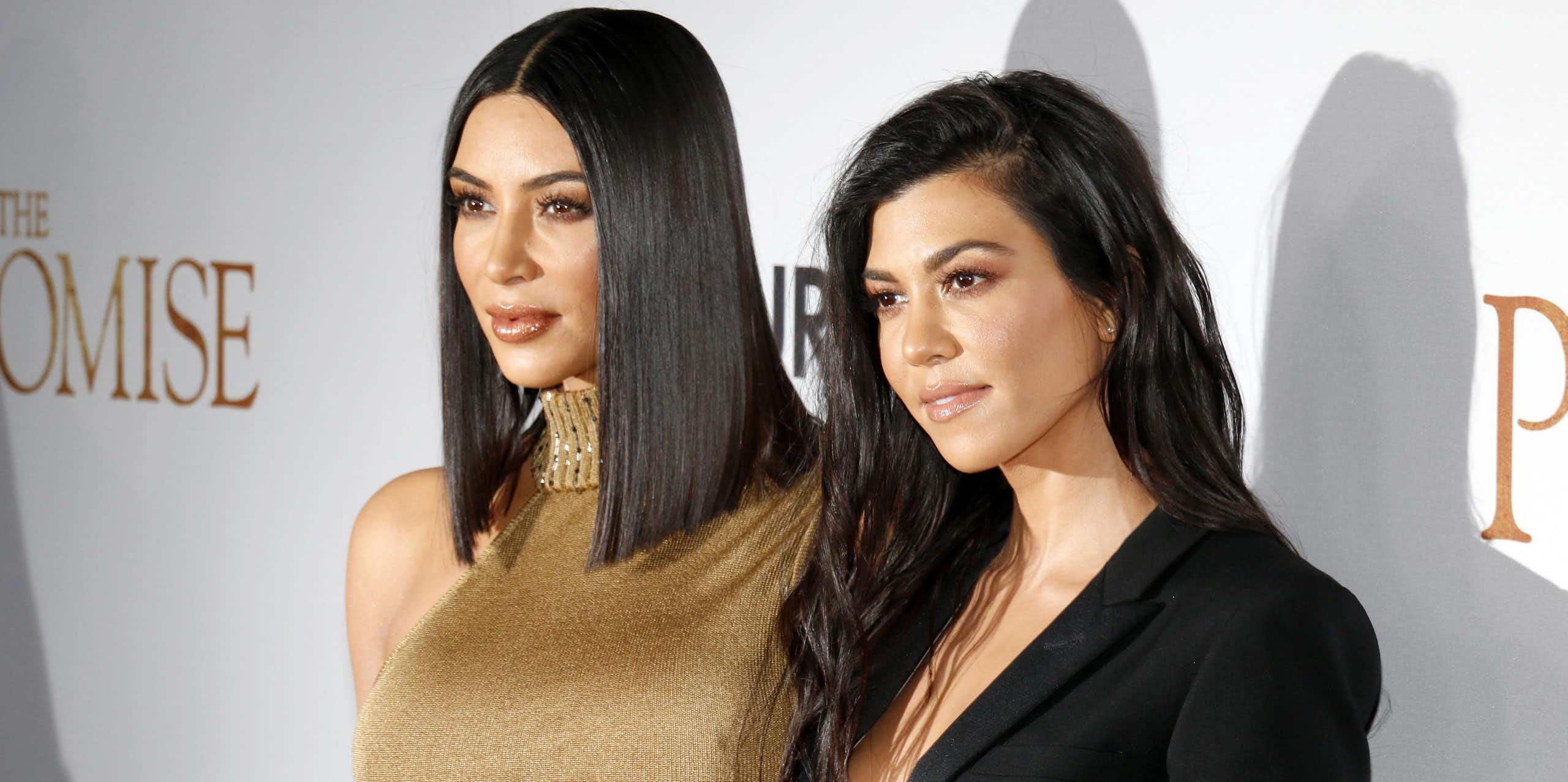 Kim Kardashian wearing a gold, sleeveless dress poses next to Kourtney Kardashian, who wears a black blazer, at an event. 