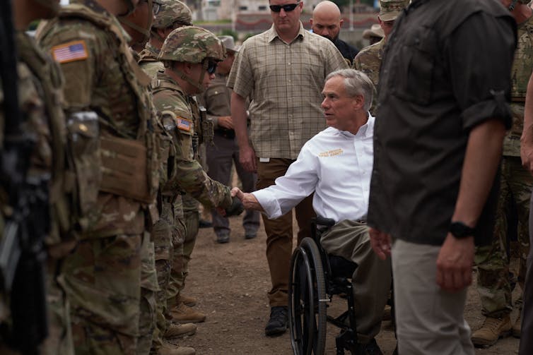 A middle aged white man in a white shirt sits in a. wheelchair and shakes the hand of a soldier who wears a camo uniform, in a row of other people in camp.