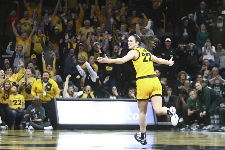 Young woman in yellow basketball uniform spreads her arms and smiles as she sprints down the court. The crowd in the background cheers wildly.