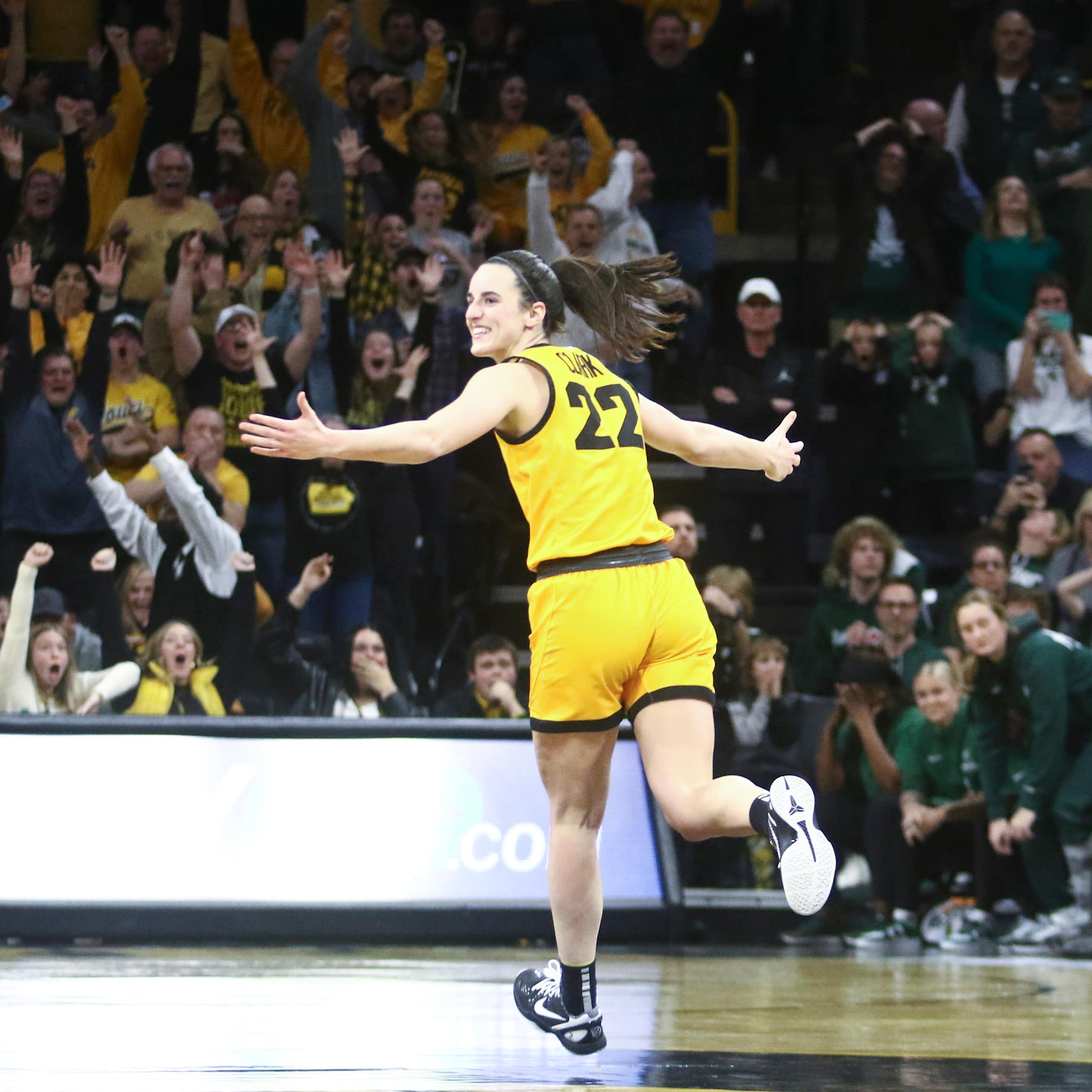 Young woman in yellow basketball uniform spreads her arms and smiles as she sprints down the court. The crowd in the background cheers wildly.