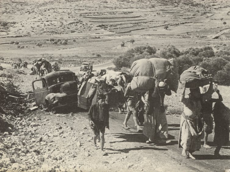 A black and white photograph of a long line of people, including women and children, walking uphill as they hold bags of possessions.
