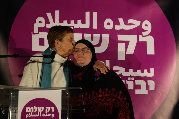 A woman in a white shirt kisses the forehead of another woman in a headscarf, whose eyes are closed, as they stand in front of a purple sign.