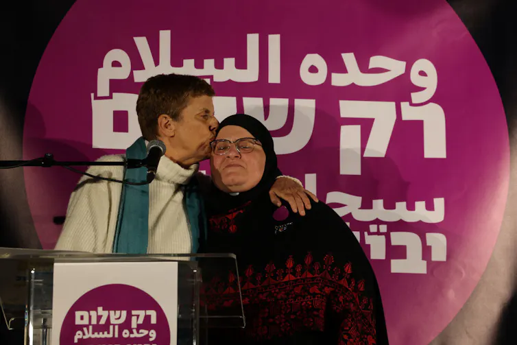 A woman in a white shirt kisses the forehead of another woman in a headscarf, whose eyes are closed, as they stand in front of a purple sign.