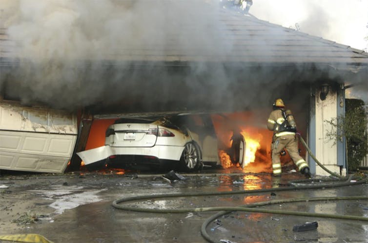 A car in a garage is on fire with the door cracked open, a firefighter carrying a hose runs towards it.