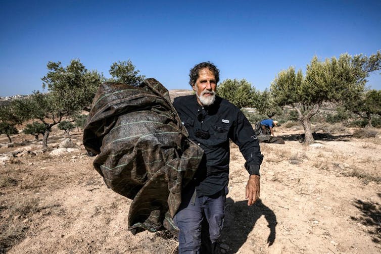 A man wearing blue, with a white beard and black hair, carries a large bundle through a dry grove of small trees.