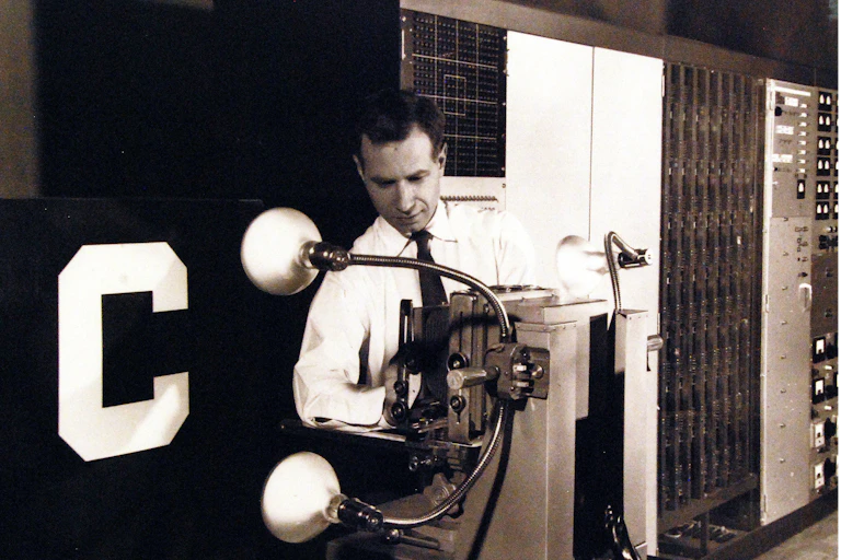 Black and white photo of a man wearing a shirt and tie standing behind a machine with two large lightbulb-like objects on flexible rods attached to its side