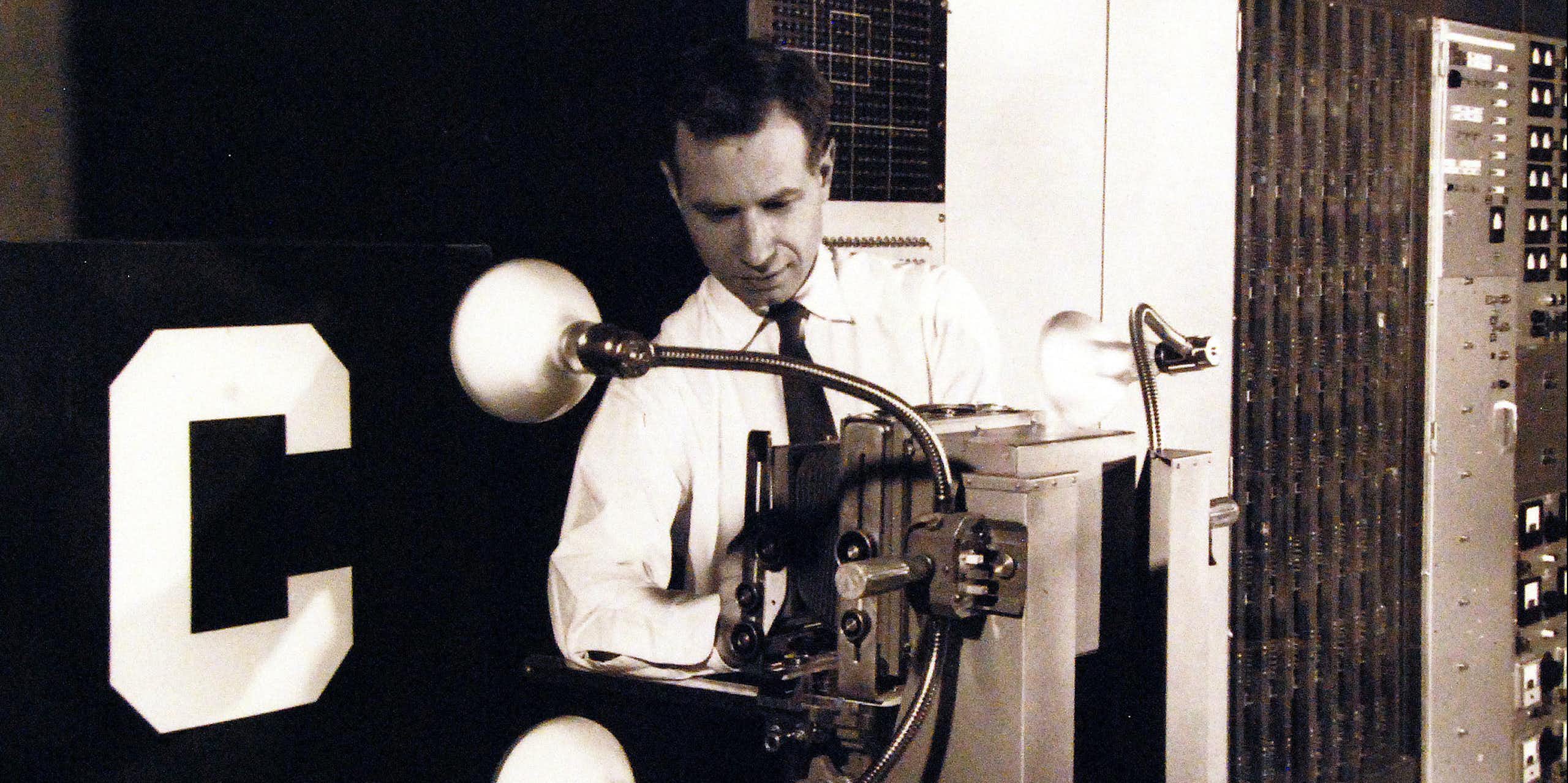 Black and white photo of a man wearing a shirt and tie standing behind a machine with two large lightbulb-like objects on flexible rods attached to its side