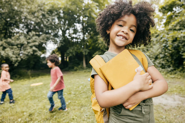 A girl with a backpack and notebook stands on a field where two boys play in the background.