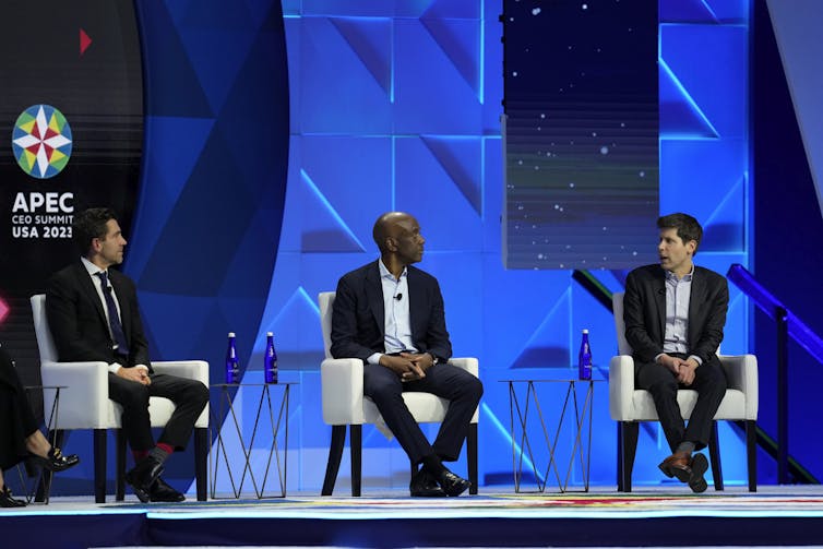 Three men sit in chairs on a stage