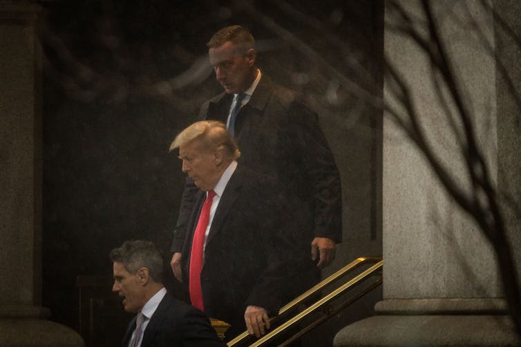 Donald Trump wears a black jacket and red tie and walks down steps outside, flanked by two men also in suits.