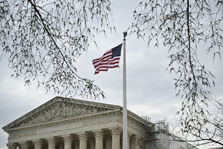 The top half of the Supreme Court building, including pillars, is seen on a gray day. The U.S. flag waves above it.