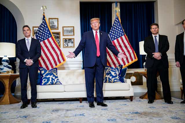 Donald Trump wears a navy blue suit and red tie and stands in front of two American flags in a room with a white couch. Two men also in navy suits stand nearby him. 