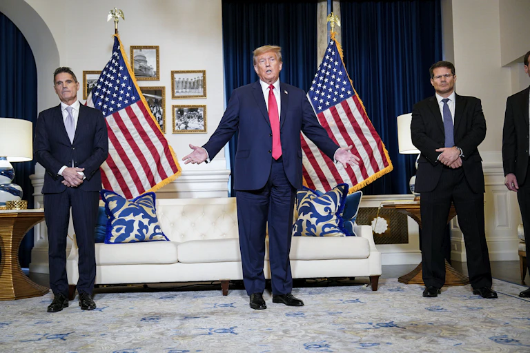 Donald Trump wears a navy blue suit and red tie and stands in front of two American flags in a room with a white couch. Two men also in navy suits stand nearby him.